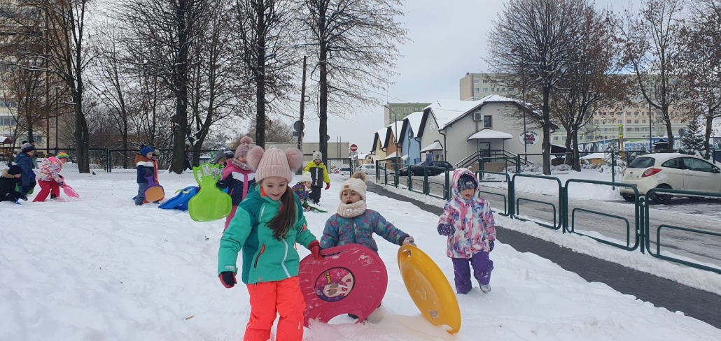 Na zdjęciu: Grupa przedszkolaków w kolorowych kombinezonach bawi się na śniegu