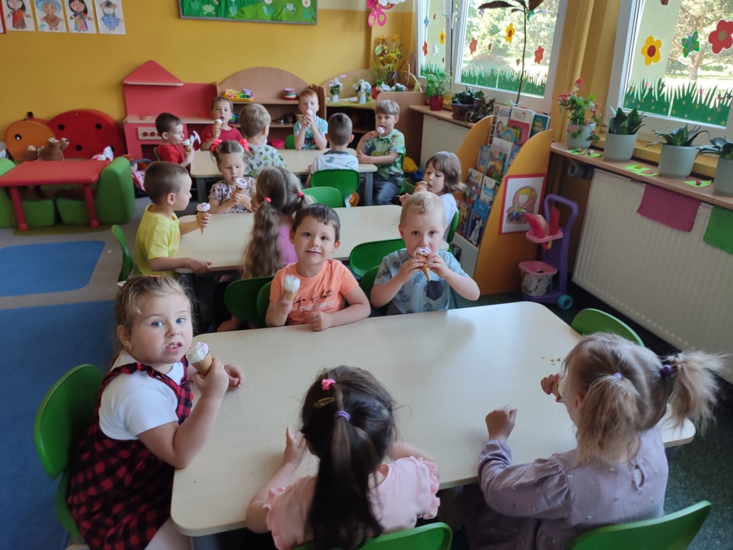 Dzieci siedzą przy stolikach i jedzą lody.