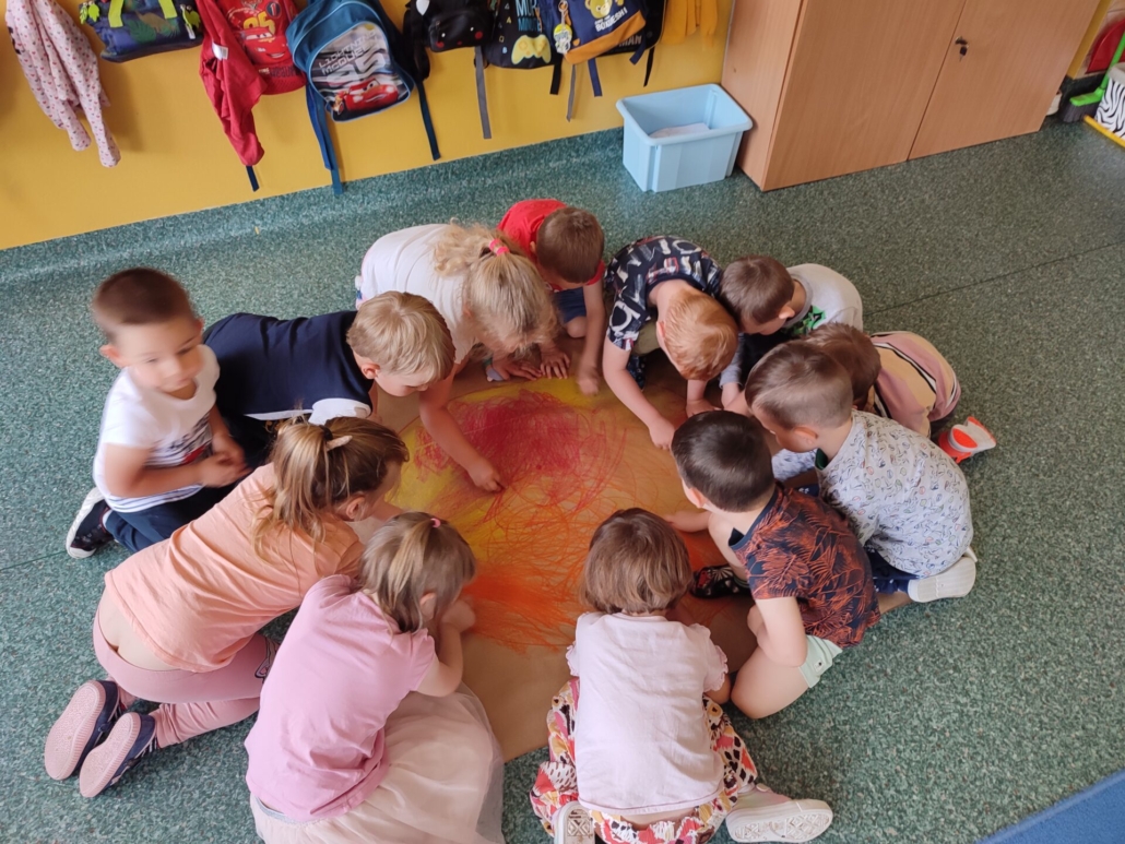  Dzieci z grupy "Motylki" są zgromadzone na podłodze wokół arkusza szarego papieru i rysują wspólnie gorące słońce.