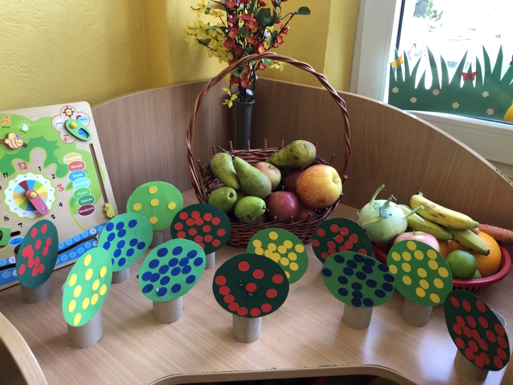 Szafka na której leżą owoce w koszykach oraz zrobione przez dzieci drzewa owocowe z kolorowego papieru i rolki po papierze toaletowym.