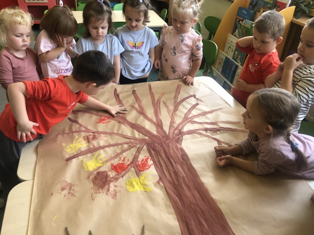 Chłopiec odbija na dużym arkuszu papieru z namalowanym drzewem swoją dłoń pomalowaną na czerwono. Dłonie zastępują jesienne liście.